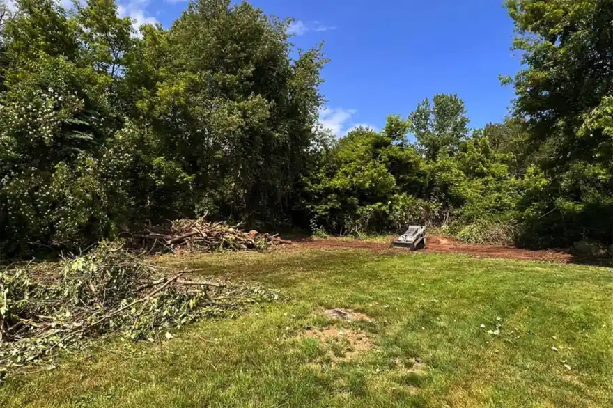 The yard during clearing, with cut brush piled at the treeline and a track loader working.