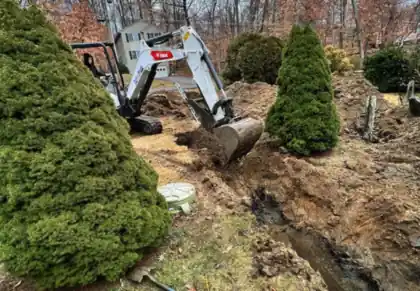 A compact excavator digging alongside foundation plantings, capped risers in the fresh soil.