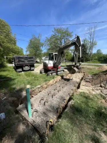 A long open trench with a green sewer line laid in it, excavator and dump truck on the lawn.