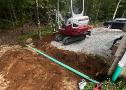 A green sewer line in an open trench with an excavator on the gravel pad above it.