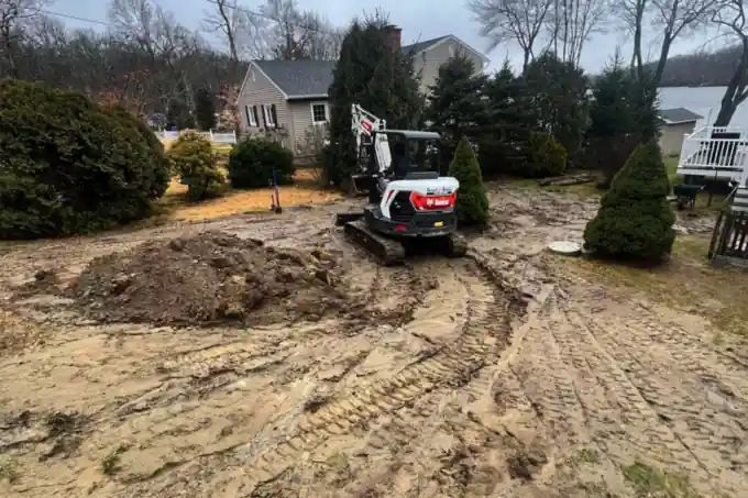 A compact excavator regrading a lakeside yard, tracked soil across the whole work area.