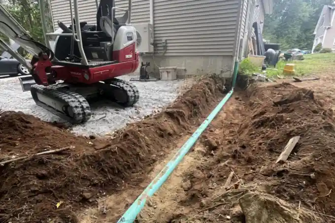 A green sewer line run in an open trench up to the side of a house, an excavator working above it.
